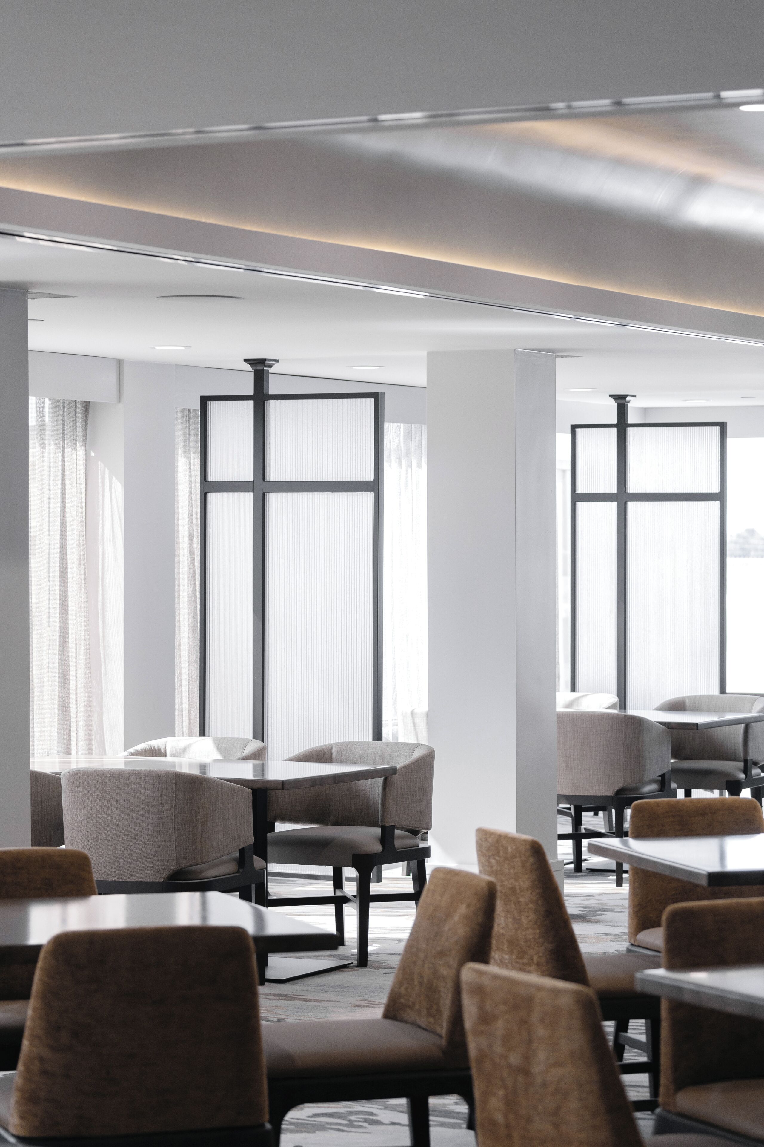 A modern hotel room with large windows, a dining area with tables and chairs, and a neutral color palette.