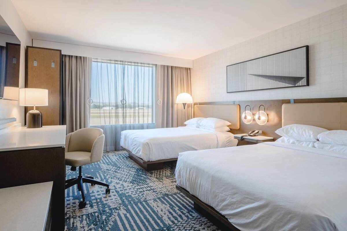A hotel room with two beds, a desk, a chair, a large window with curtains, and a wall-mounted artwork.
