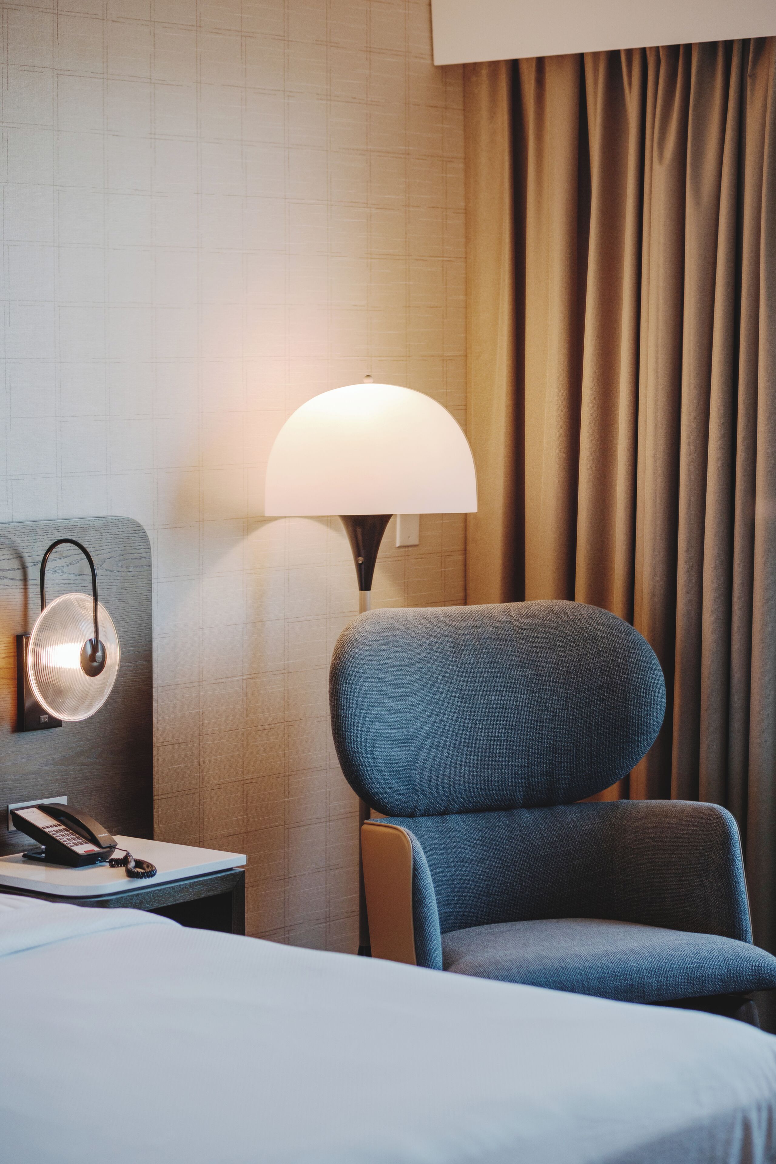 Quarto de hotel com cama, cadeira, abajur, telefone e cortinas.