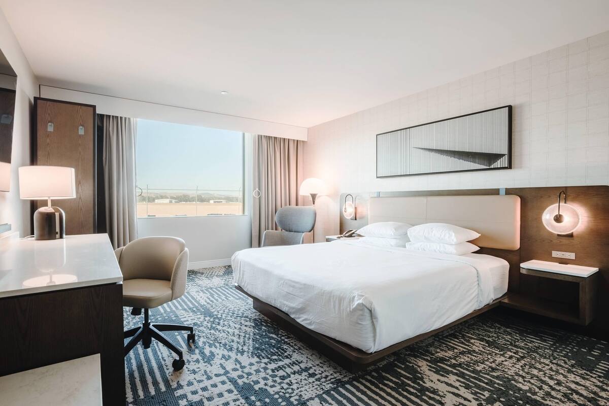A hotel room with a bed, desk, chair, bedside lamps, and a large window.