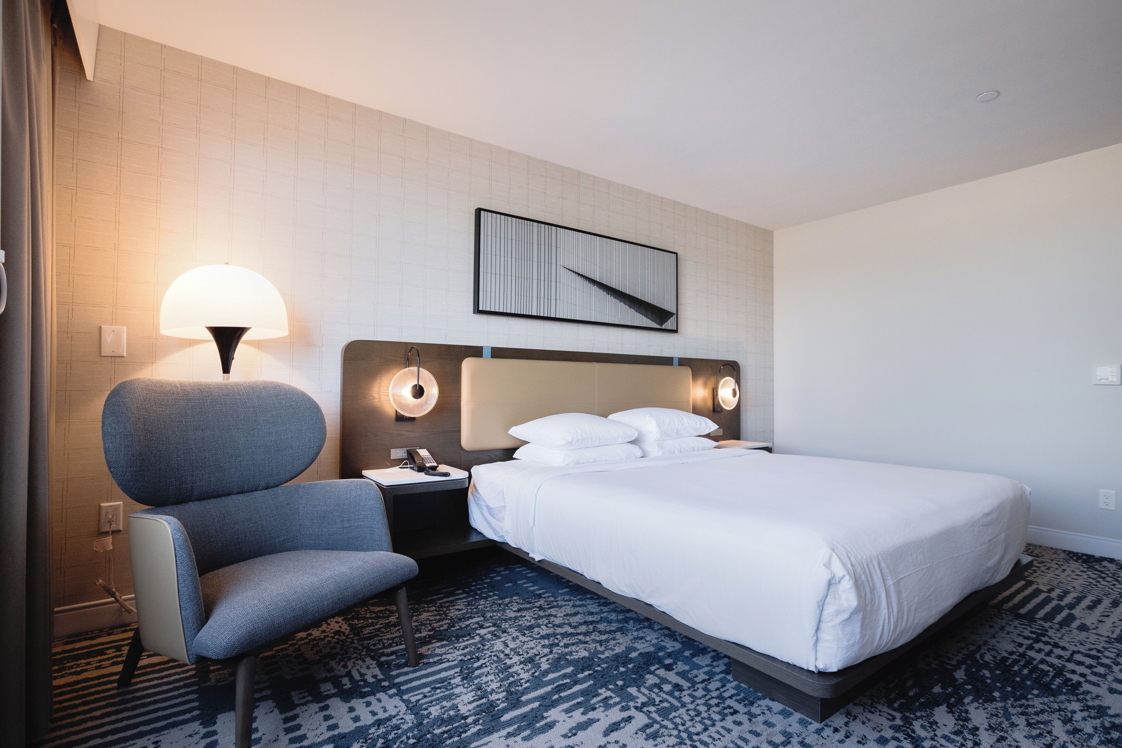 Una habitación de hotel con cama, una silla, una mesita de noche, una lámpara y una obra de arte colgada en la pared.