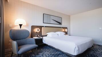 Una habitación de hotel con cama, una silla, una mesita de noche, una lámpara y una obra de arte colgada en la pared.
