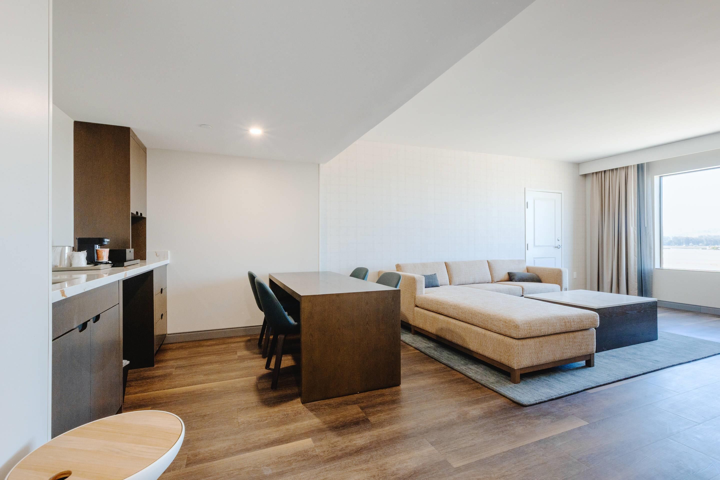 Een moderne hotelkamer met een houten vloer, een eettafel met stoelen, een bank, een kleine keukenhoek en een groot raam.