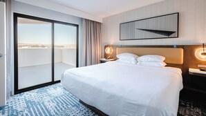 A hotel room with a large bed, a sliding glass door to a balcony, a wall-mounted artwork, and a bedside table with a lamp.