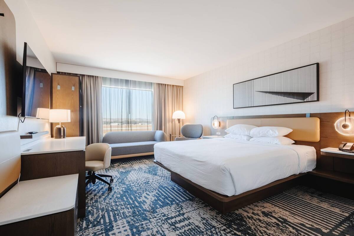A hotel room with a bed, desk, chair, and a large window with curtains.