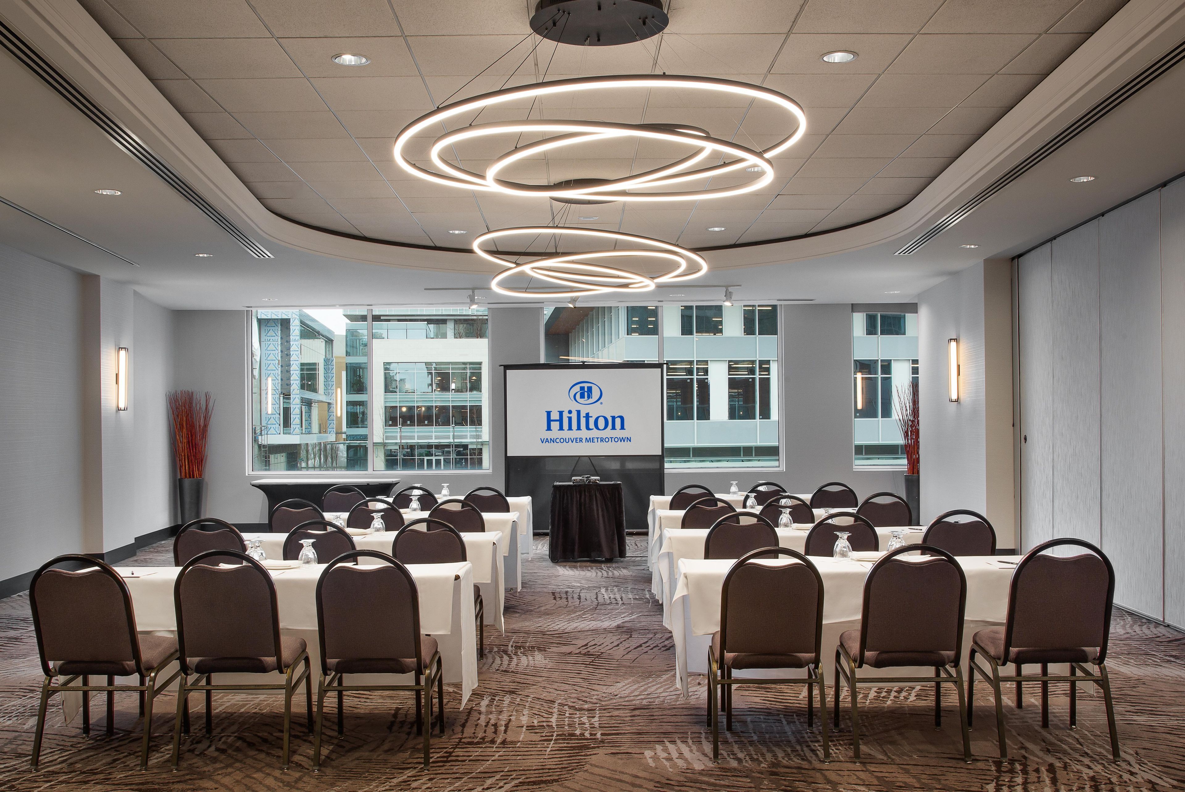 Ein Konferenzraum mit Tischen und Stühlen, einem großen Bildschirm mit dem Hilton-Logo und einem modernen Deckenstil.
