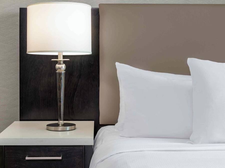 A neatly made bed with white linens and a single pillow, a bedside table with a lamp, and a dark-colored nightstand.