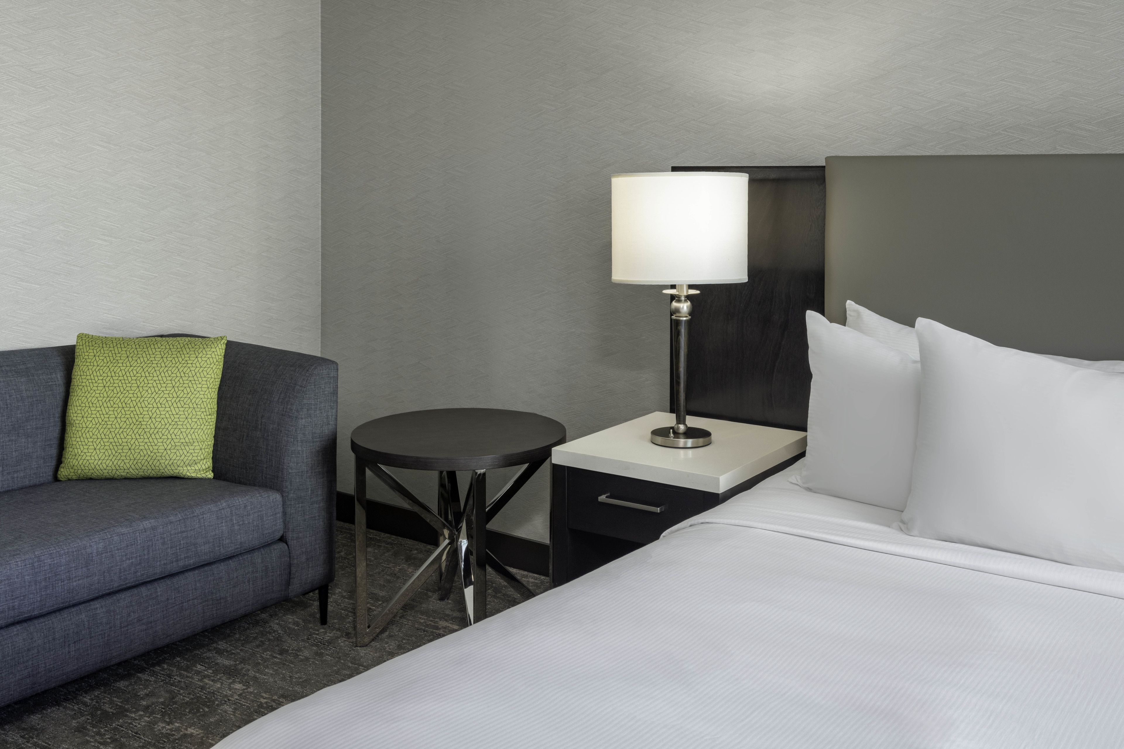 Una habitación de hotel con cama, mesita de noche, lámpara, una pequeña mesa y un sofá con almohada.
