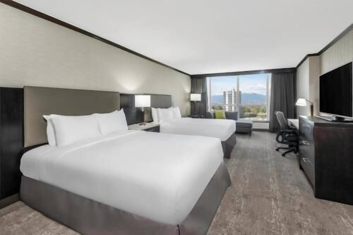 Ein Hotelzimmer mit zwei Betten, einem Schreibtisch, einem Stuhl, einem Fernseher und einem großen Fenster mit Stadtblick.