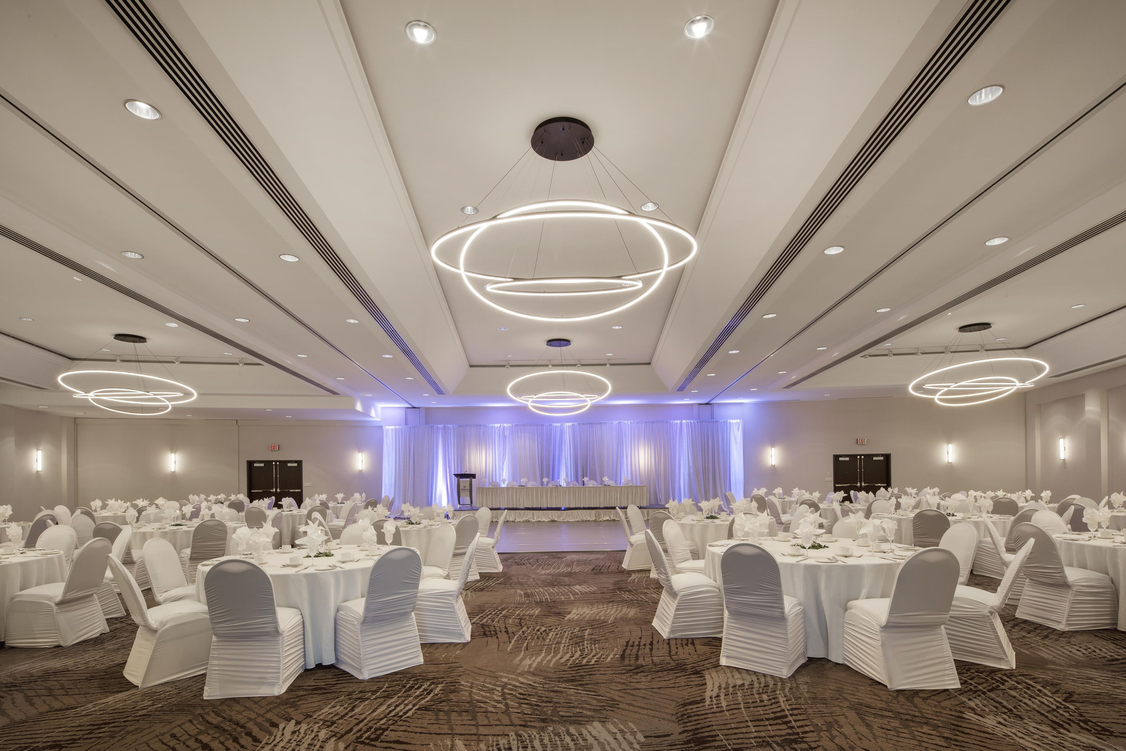 Un salón de banquetes con mesas redondas, sillas blancas y un escenario con cortinas.