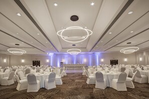 Un salón de banquetes con mesas redondas, sillas blancas y un escenario con cortinas.