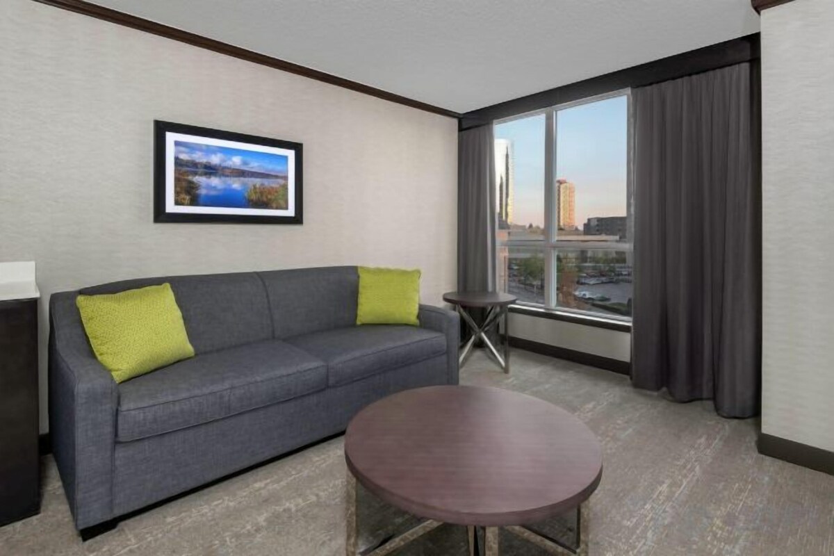A hotel room with a gray sofa, a small round table, a framed picture on the wall, and a large window with curtains.
