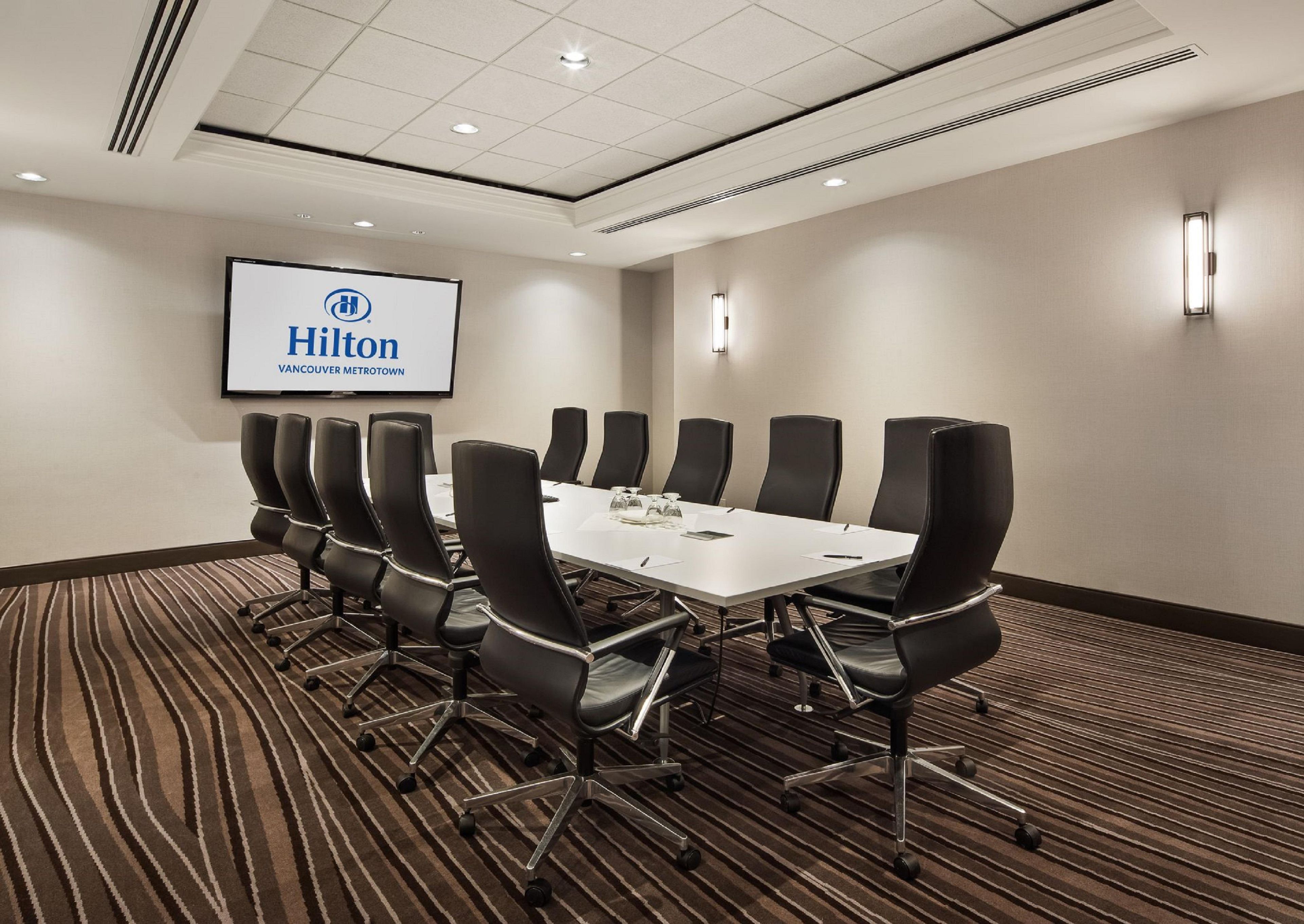 A conference room with a long table and chairs, a screen displaying 'Hilton Vancouver Metrotown', and a carpeted floor.