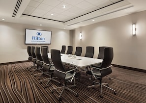 Un salón de conferencias con una larga mesa y sillas, una pantalla mostrando 'Hilton Vancouver Metrotown' y un piso alfombrado.