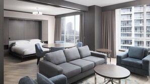 Habitación de hotel moderna con una cama grande, sofá gris, sillones, una mesita y vistas a la ciudad a través de amplios ventanales.