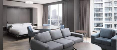 Habitación de hotel moderna con una cama grande, sofá gris, sillones, una mesita y vistas a la ciudad a través de amplios ventanales.
