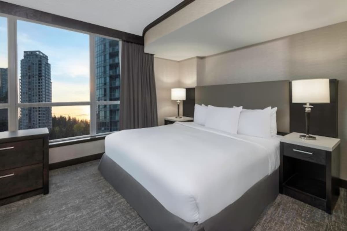Una habitación de hotel con una cama grande, dos mesitas de noche, un ventanal con vistas a edificios y un paisaje urbano.