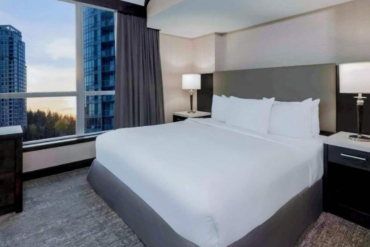 Ein Hotelzimmer mit einem großen Bett, zwei Nachttischen, einem Fenster mit Blick auf Gebäude und einer Stadtansicht.
