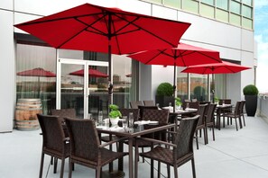 Mesas de comedor al aire libre con sillas de mimbre y sombrillas rojas, frente a un edificio con grandes ventanales.