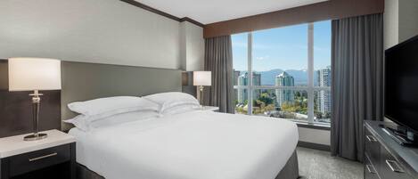 Une chambre d’hôtel avec un grand lit, deux tables de chevet avec des lampes, un téléviseur à écran plat et une vue sur la ville grâce à de grandes fenêtres.