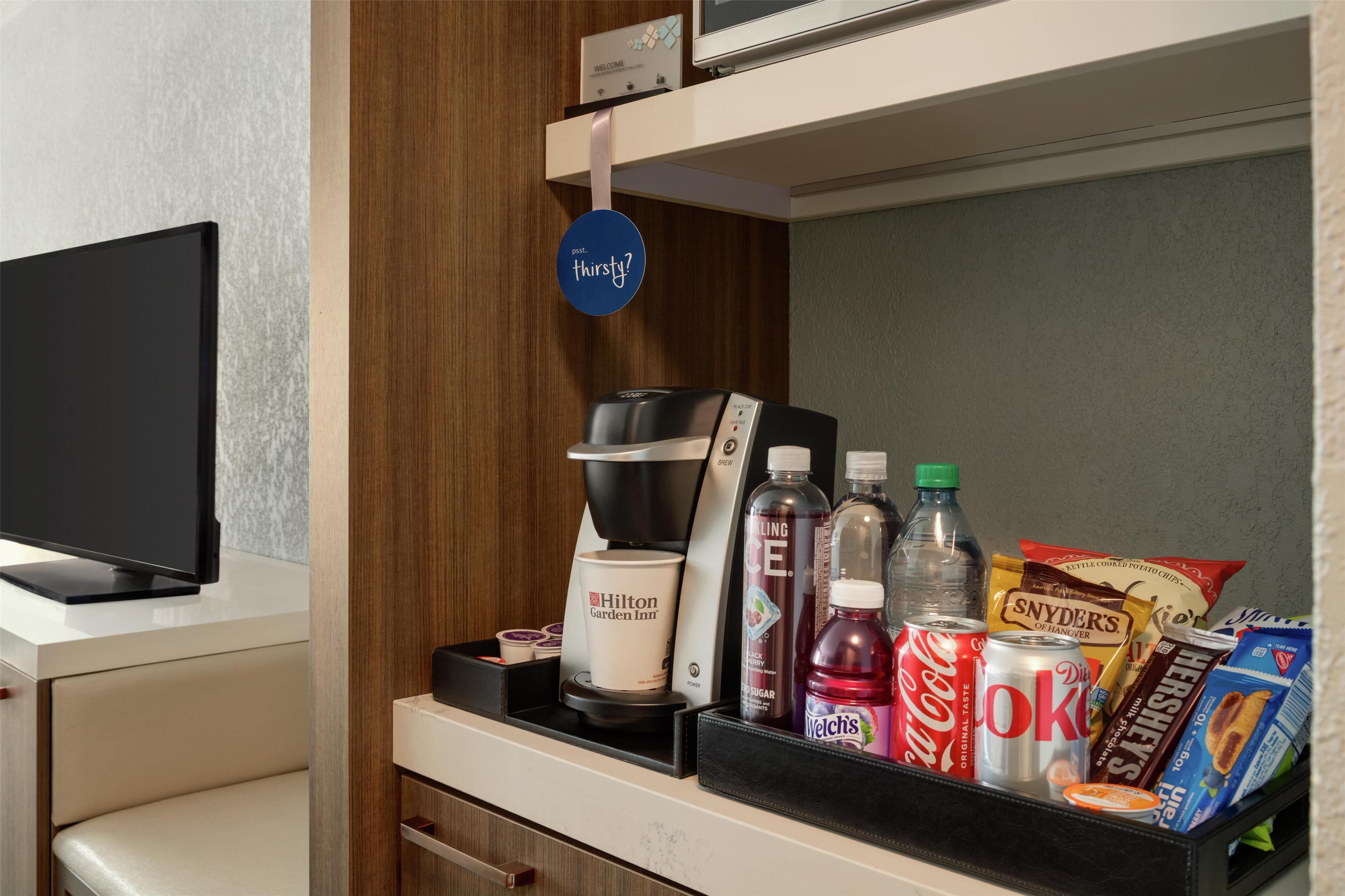 Un minibar con una cafetera, una botella de Coca-Cola, una lata de Diet Coke, una botella de Welch's, una bolsa de Reese's y una caja de detergente Tide.