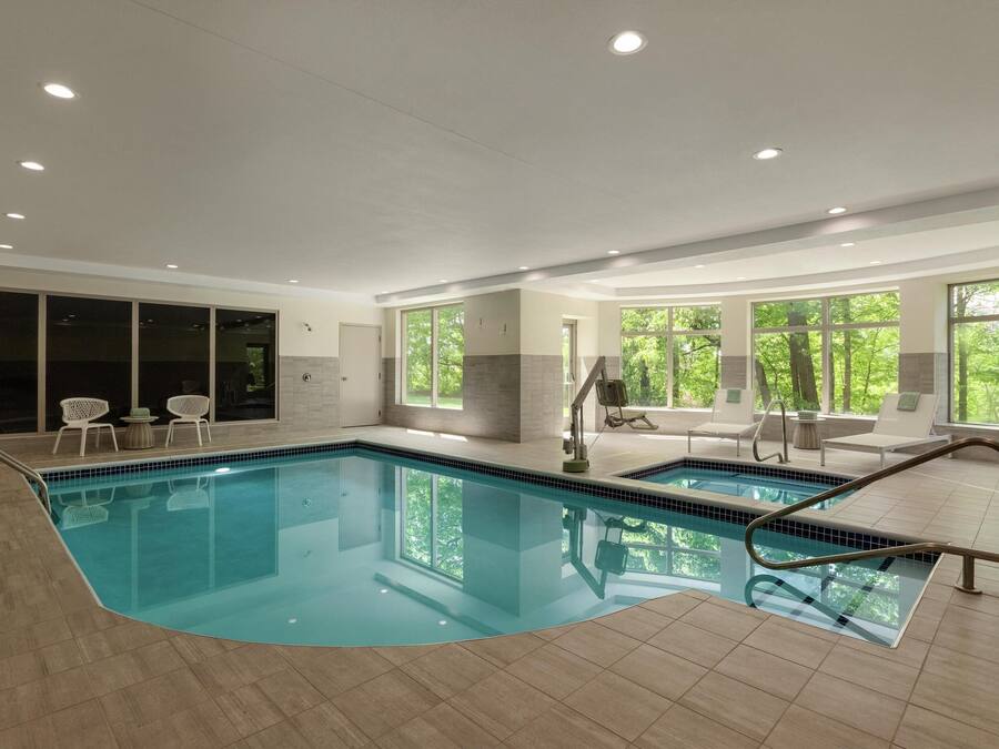 Piscina interior com paredes de vidro transparente, área ensolarada com assentos e vista para o verde lá fora.