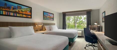 A hotel room with two beds, a desk, a chair, a TV, and large windows with a view of trees.