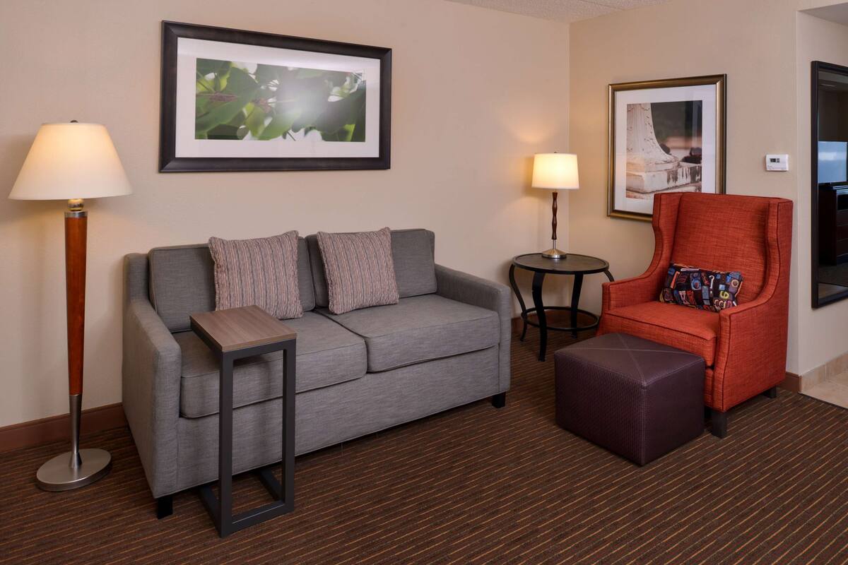 Ein Hotelzimmer mit grauem Sofa, einem orangefarbenen Sessel, einem kleinen runden Tisch und zwei Lampen.