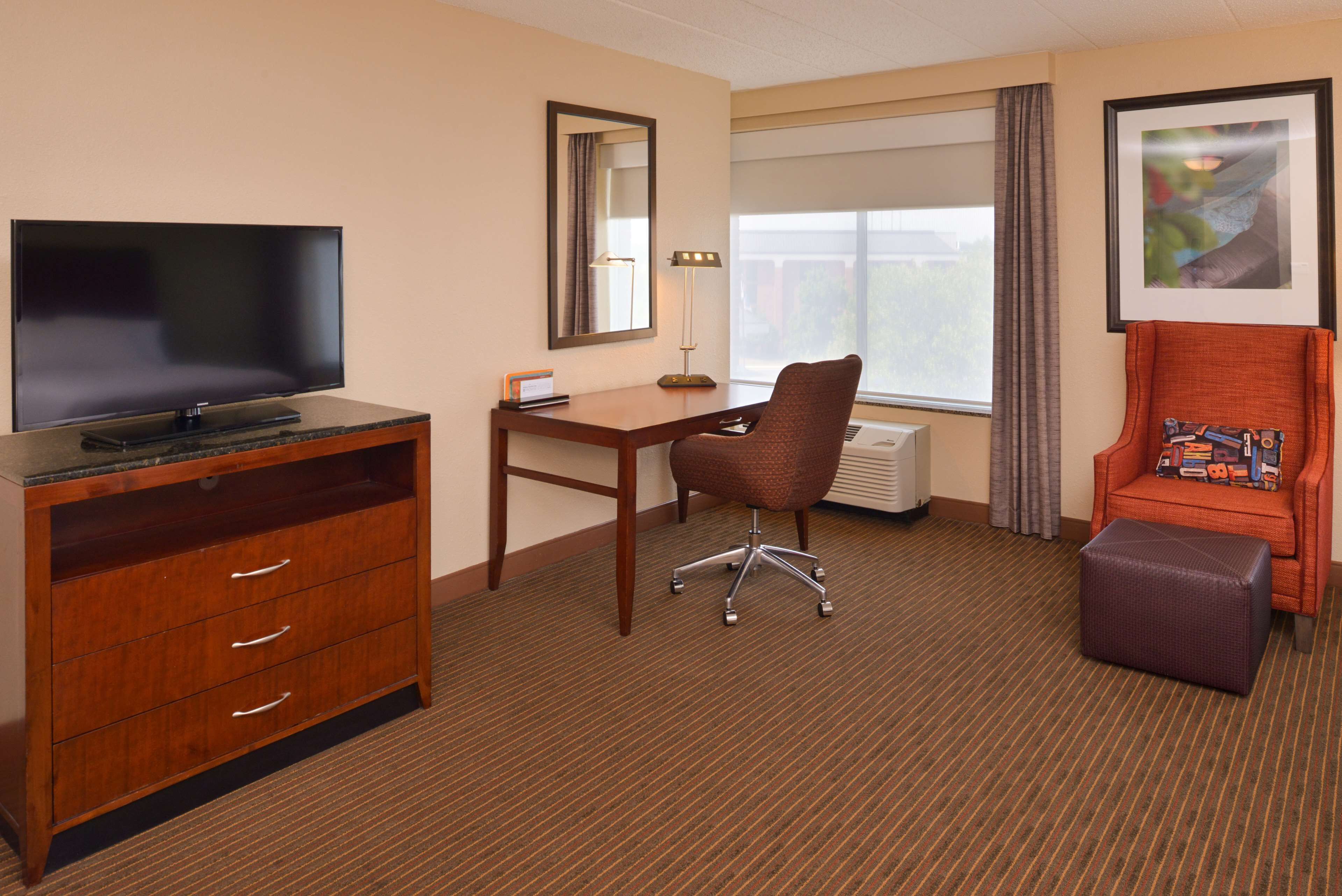 A hotel room with a flat-screen TV, a desk, a chair, a small ottoman, and a framed picture on the wall.