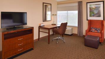 A hotel room with a flat-screen TV, a desk, a chair, a small ottoman, and a framed picture on the wall.