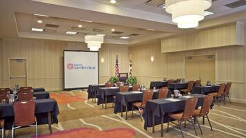 Una sala de conferencias con mesas y sillas, una pantalla de proyector mostrando 'Hilton Garden Inn' y dos banderas.
