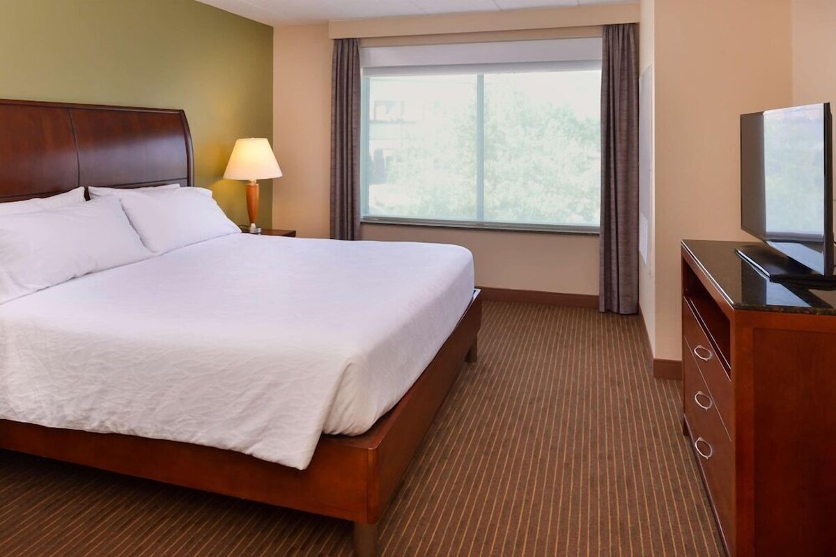 A hotel room with a large bed, a nightstand with a lamp, a television, and a window with curtains.