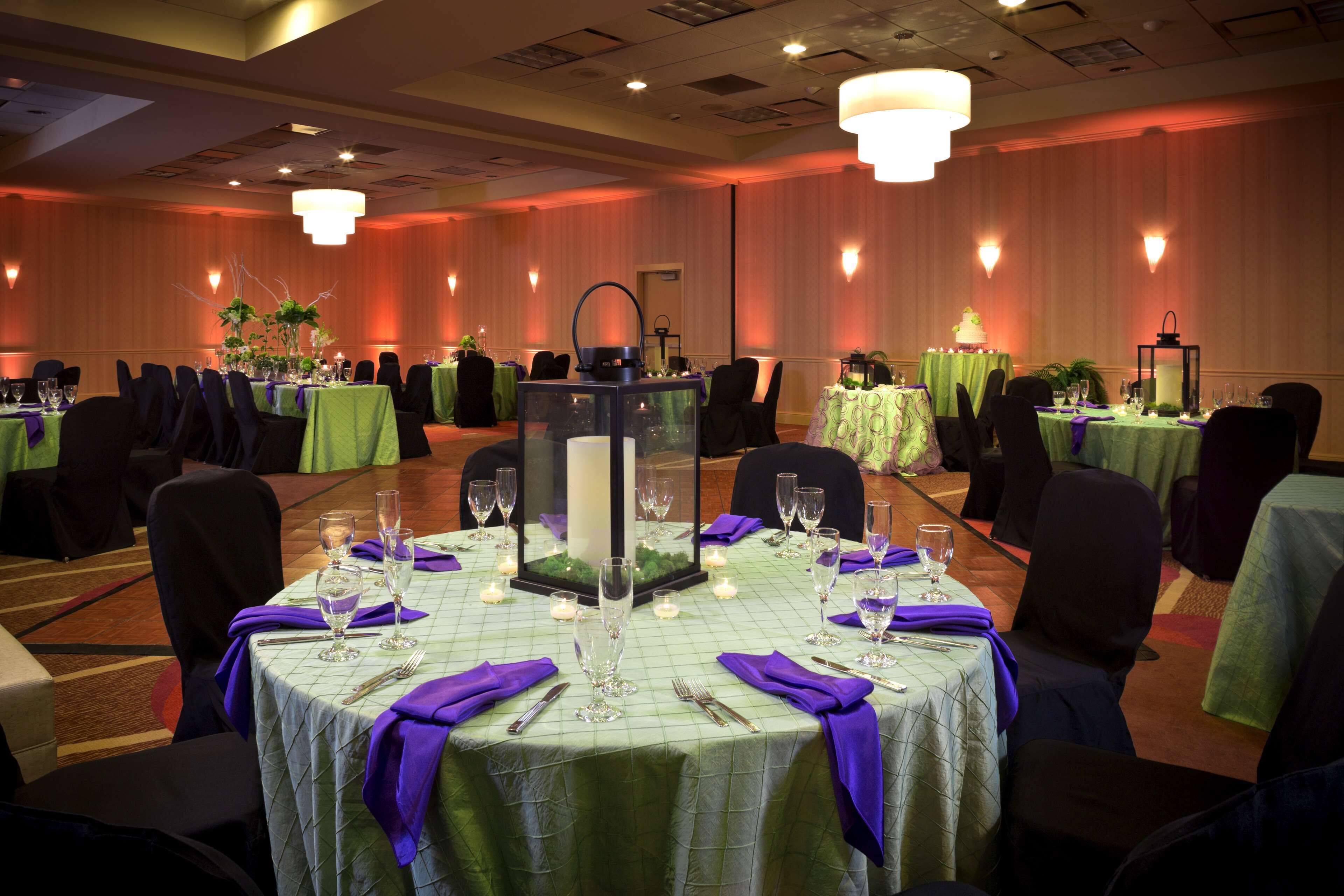 Une salle de banquet où les tables sont dressées pour un événement formel, avec des nappes vertes, des serviettes violettes et des centres de table ornés de bougies.