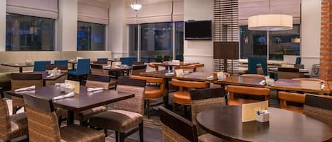 Un comedor de hotel con varias mesas y sillas, ventanas amplias y un televisor de pantalla plana montado en la pared.