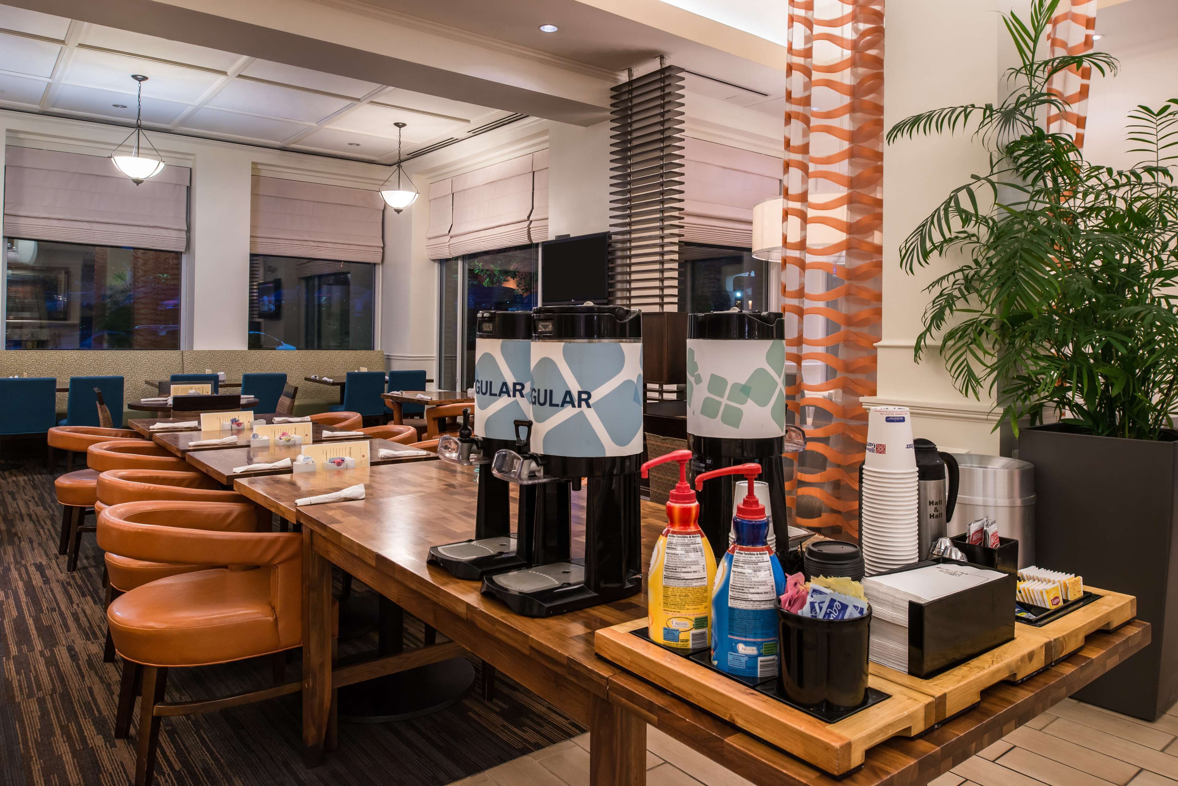 Un espace dédié au petit-déjeuner dans l’hôtel, avec une longue table en bois, de nombreuses chaises, des machines à café et une variété d’aliments proposés pour le petit-déjeuner.