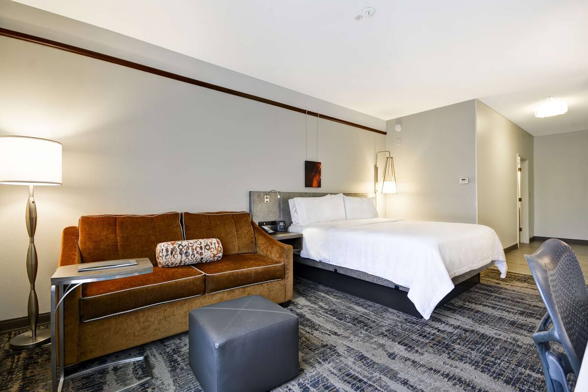 A hotel room with a brown sofa, a bed with white linens, a gray ottoman, a standing lamp, and a modern floor lamp.