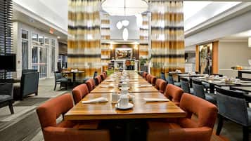 A spacious hotel dining area with long tables, comfortable seating, and a central chandelier.