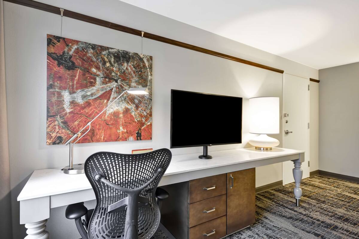 A hotel room with a desk, a chair, a flat-screen TV, a lamp, and a large abstract painting on the wall.