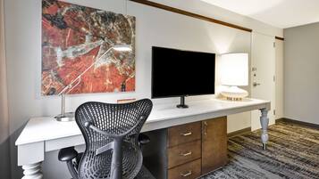 A hotel room with a desk, a chair, a flat-screen TV, a lamp, and a large abstract painting on the wall.