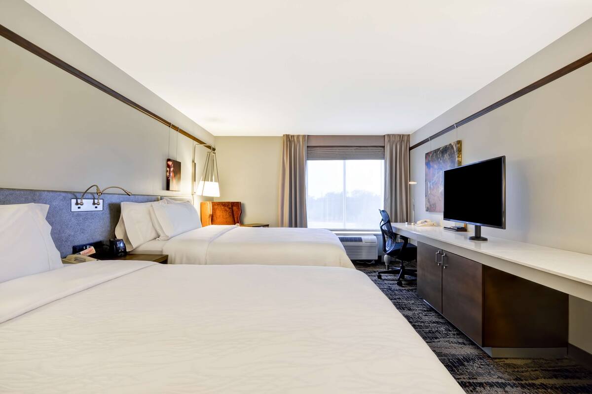 A hotel room with two beds, a desk with a flat-screen TV, a desk lamp, and a window with curtains.
