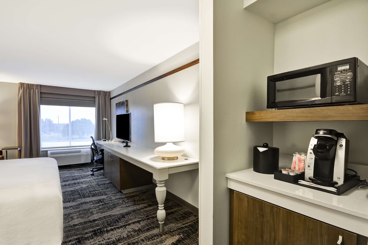 A hotel room with a bed, desk, lamp, microwave, coffee maker, and a view of the city.