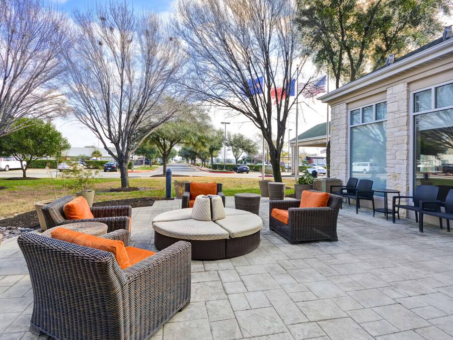 An outdoor patio with wicker furniture and a stone floor, surrounded by trees and a building with large windows.