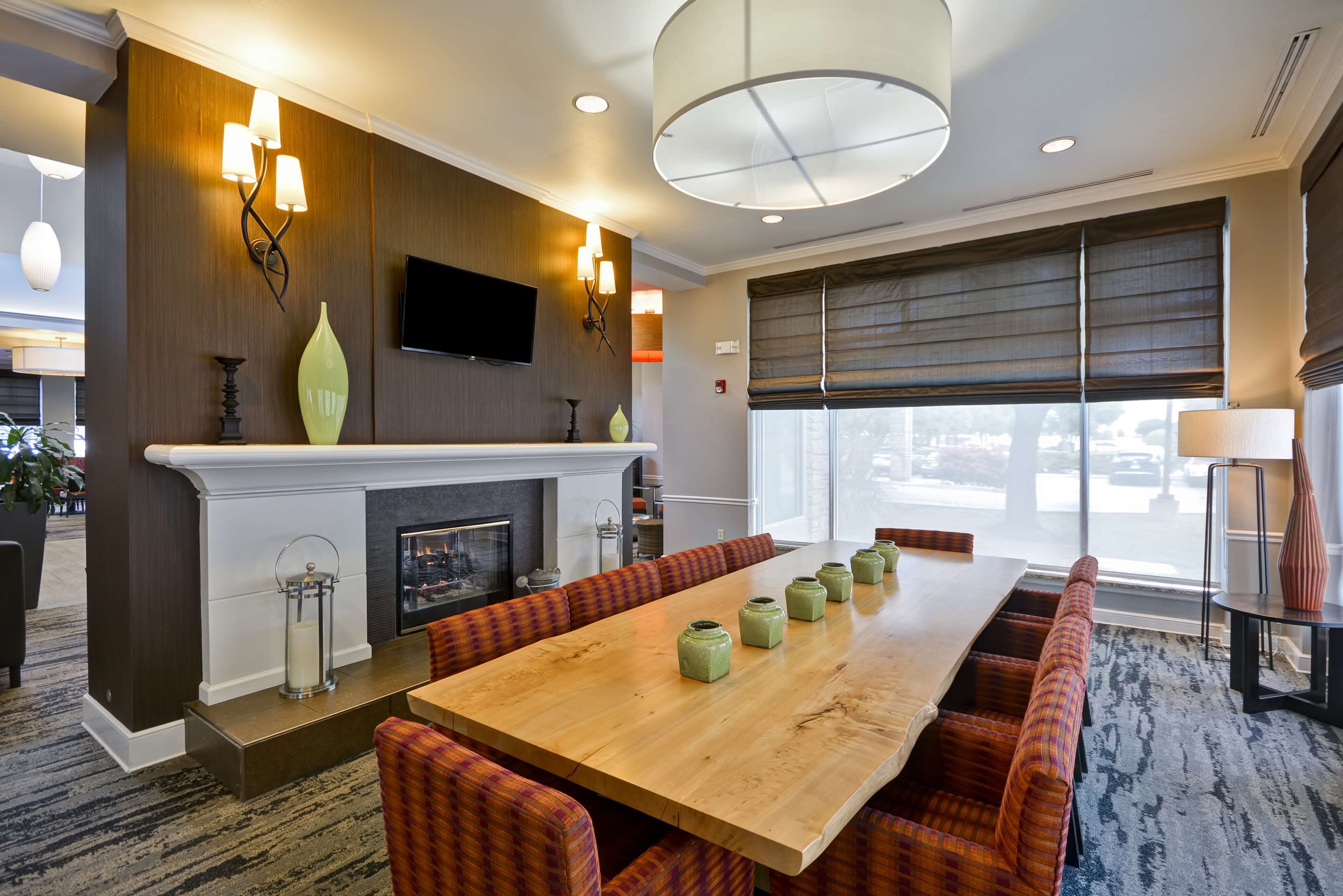 A hotel dining area with a long wooden table, cushioned benches, a fireplace, and a large window with blinds.
