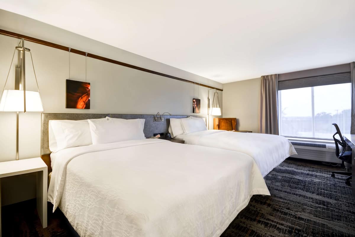 A hotel room with two beds, a desk, and a chair. There are two wall-mounted lamps and a window with curtains.