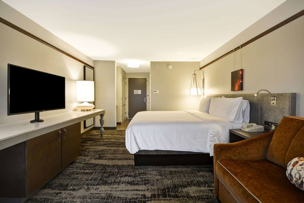 A hotel room with a bed, a desk with a TV, a chair, a bedside table with a lamp, and a painting on the wall.