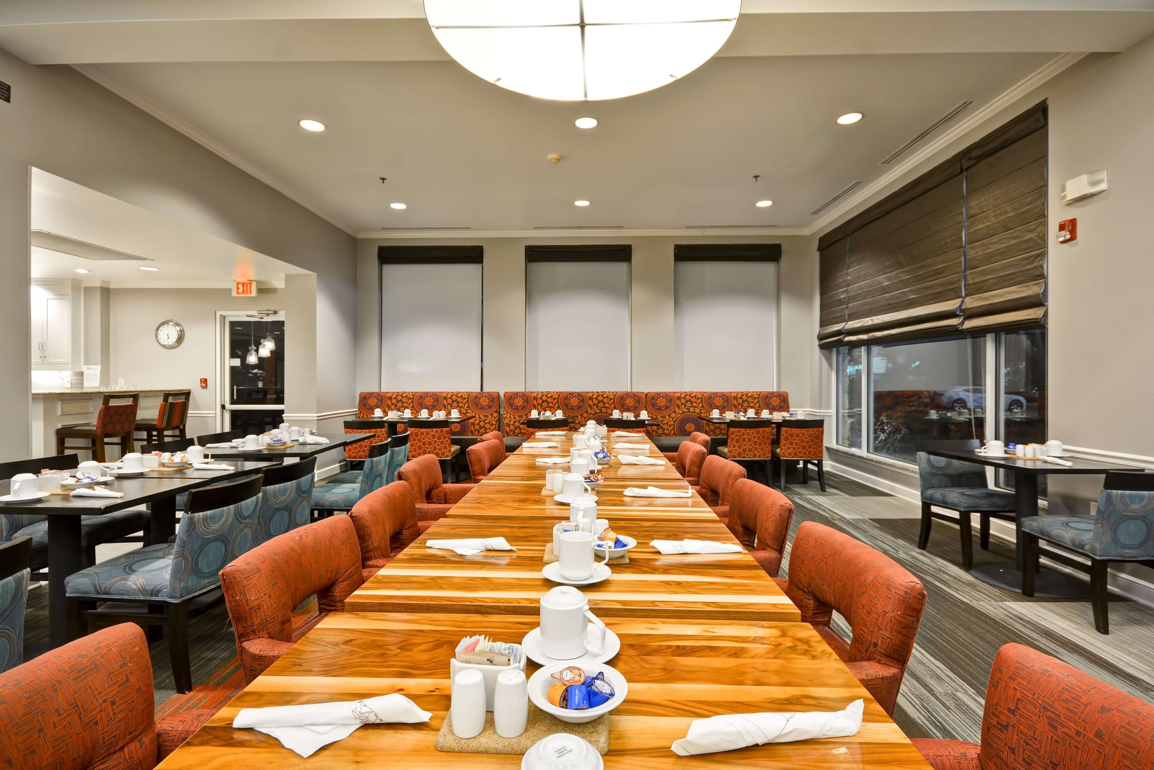 A conference room with a long table set for a meeting, surrounded by chairs, and a kitchen area visible in the background.