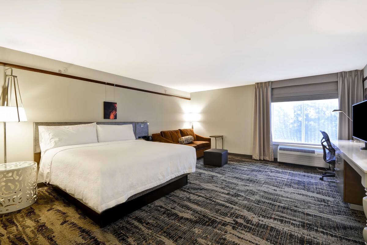 A hotel room with a large bed, a sofa, a desk, and a chair. There is a window with curtains, a lamp, and a picture on the wall.