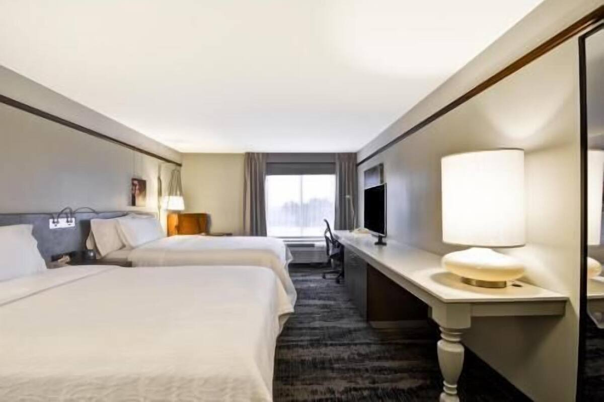A hotel room with two beds, a desk, a TV, and a window.