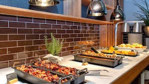 A buffet with various dishes, including meats and vegetables, in a modern kitchen setting with brick walls and wooden shelves. - Holiday Inn Berlin Airport - Conference Centre by IHG (Schoenefeld)
