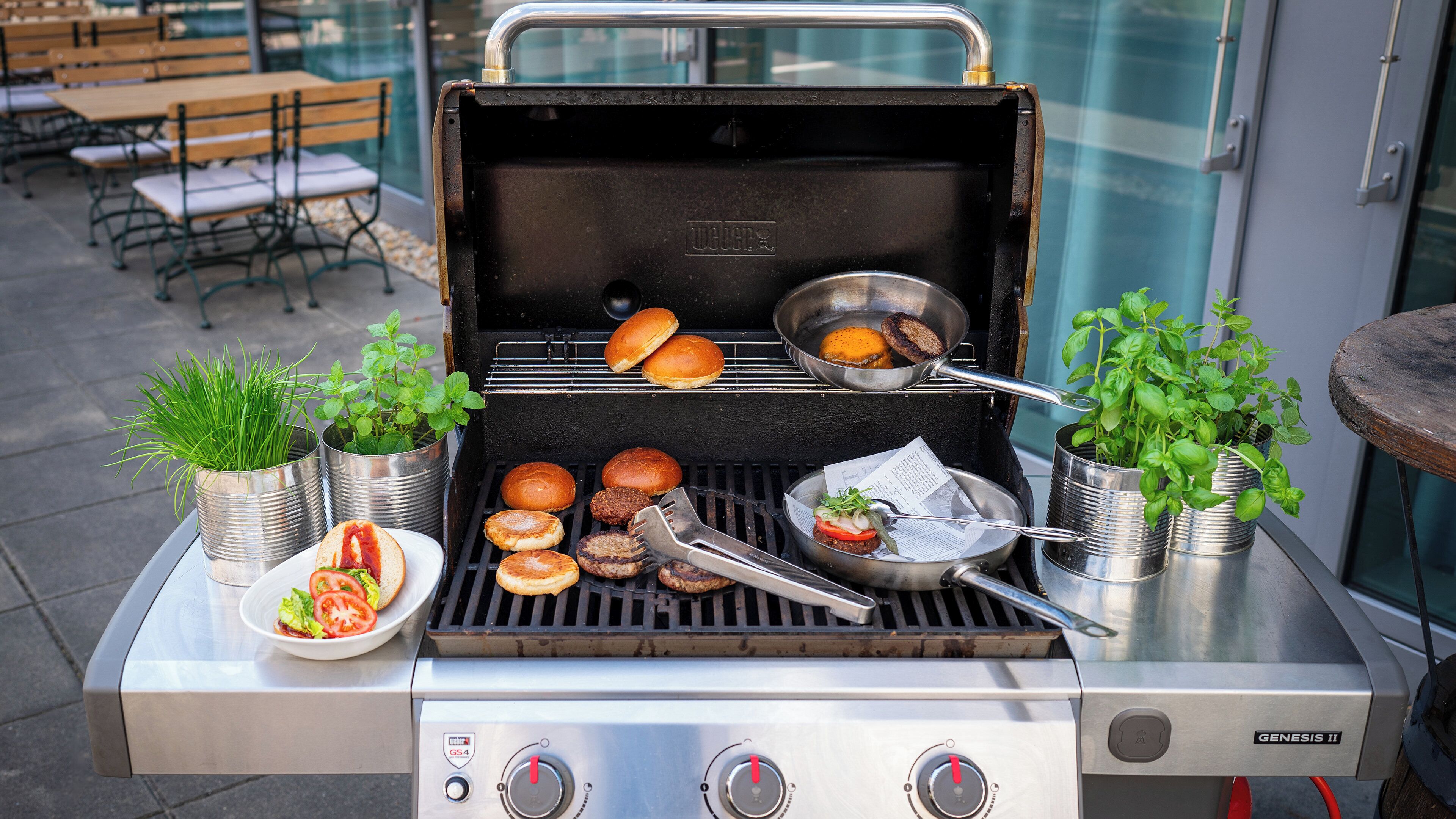 A Genesis II portable grill with multiple cooking zones, a stainless steel table, and potted plants.